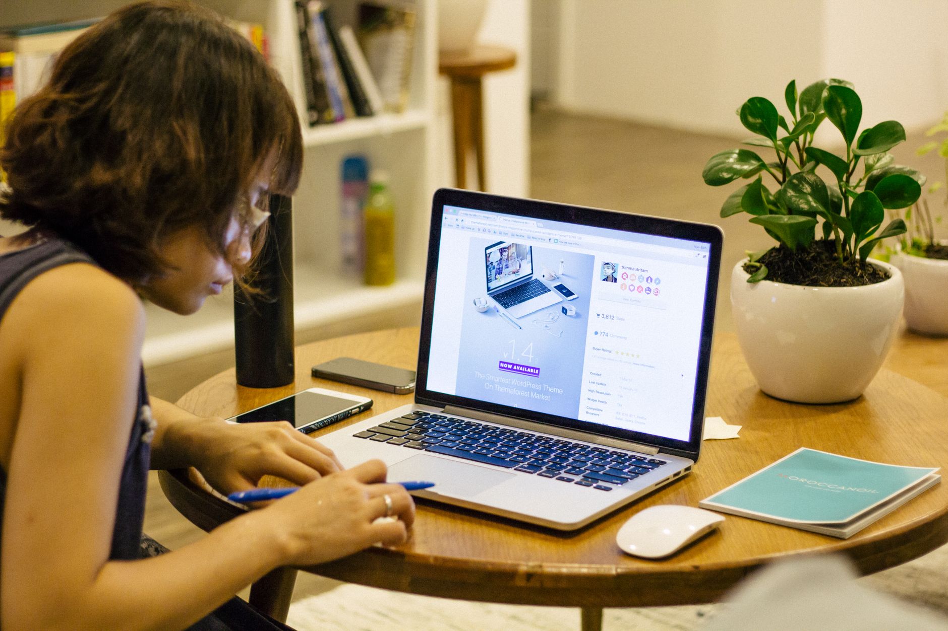 Femme travaillant à distance depuis chez elle avec un ordinateur portable, un stylo et une plante sur son bureau, illustrant le mode de vie du travail depuis la maison.
