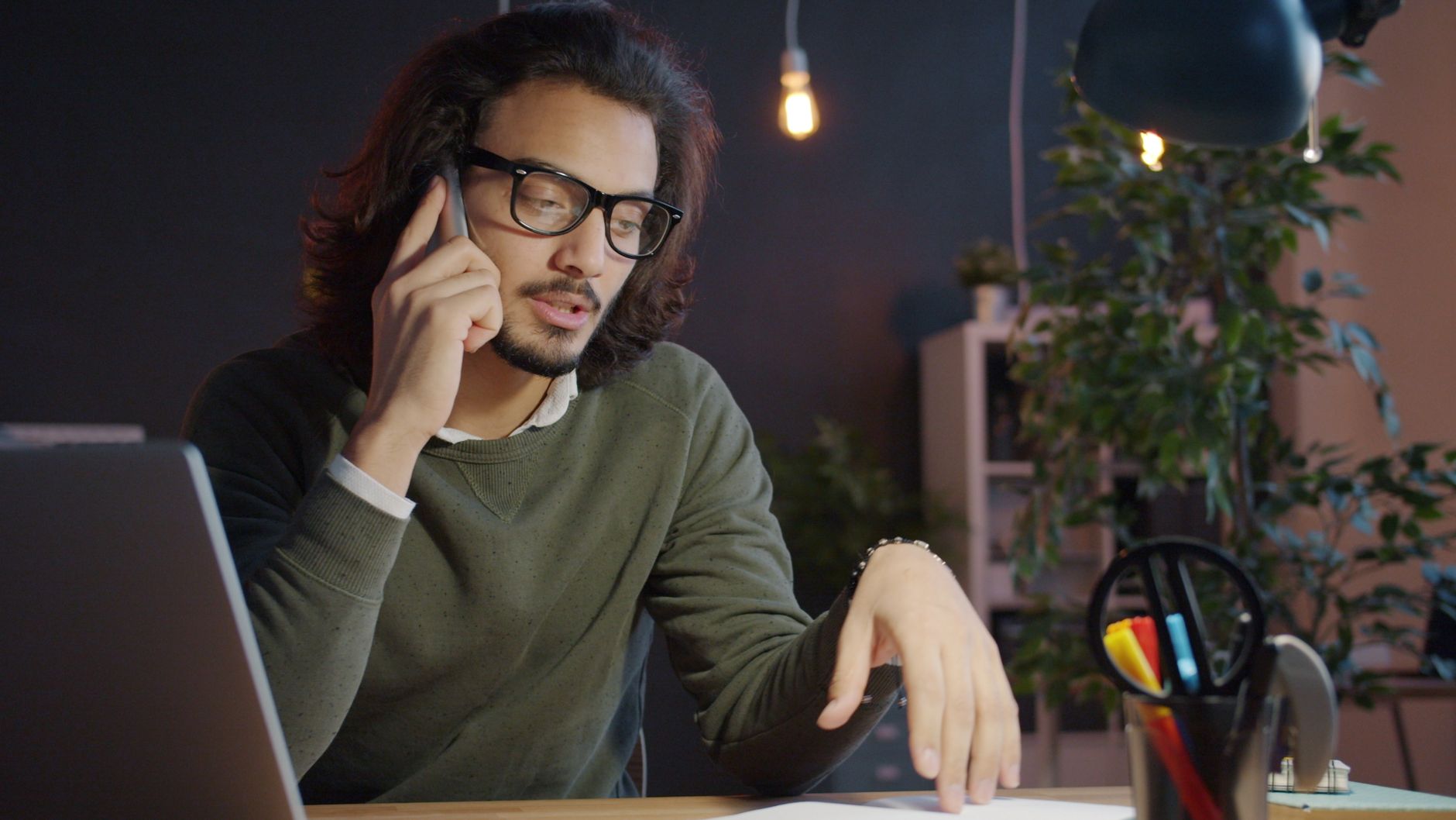 Homme adulte avec des lunettes au téléphone à son bureau de bureau à domicile avec ordinateur portable et plantes.